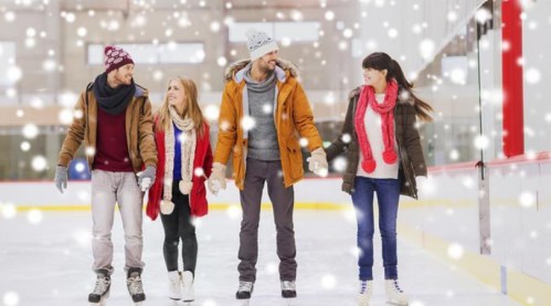 Four friends ice skating together in a snowy indoor rink, smiling and holding hands, dressed in winter attire.