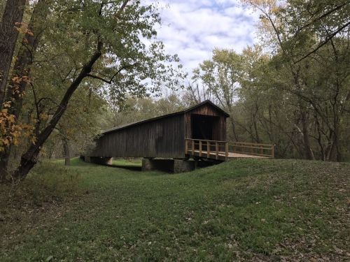 A rustic covered bridge surrounded by trees and greenery under a cloudy sky.