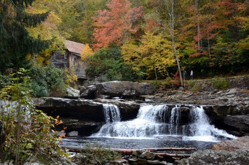A serene waterfall flows past rocks, with a historic building and vibrant autumn foliage in the background.