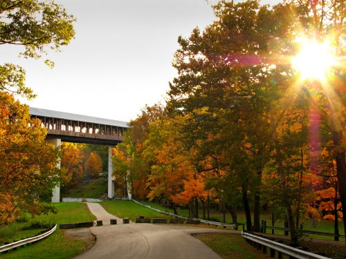 A scenic view of a covered bridge surrounded by vibrant autumn trees and a sunburst in the background.