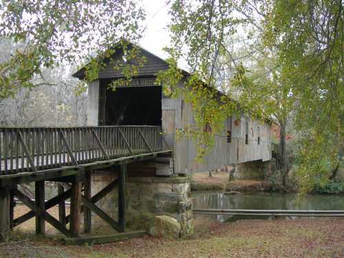 A rustic covered bridge with a wooden walkway, surrounded by trees and reflecting in a calm pond.