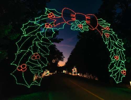A colorful holiday light display featuring a holly archway over a dark road at dusk.