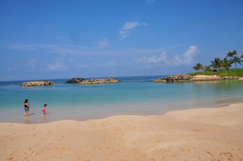 Two people walk along a sandy beach, with calm turquoise water and rocky formations in the background under a clear blue sky.