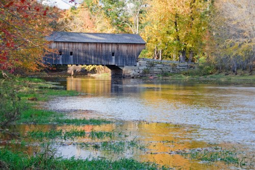 A wooden covered bridge spans a calm river, surrounded by colorful autumn foliage and reflections in the water.