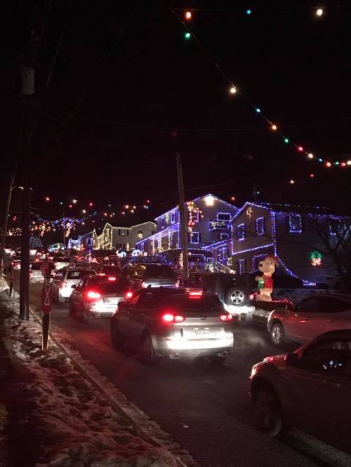 A street lined with cars and houses decorated with colorful Christmas lights at night. Snow on the ground.