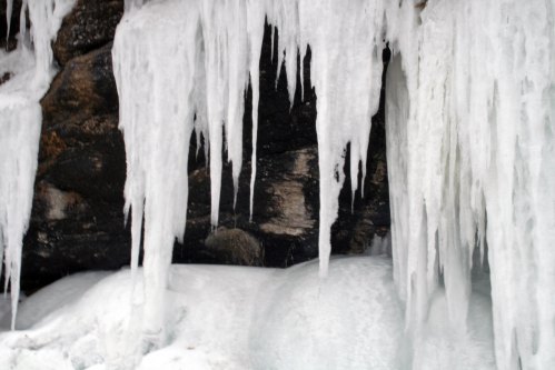 Icicles hang from a rocky surface, with a layer of snow covering the ground below.