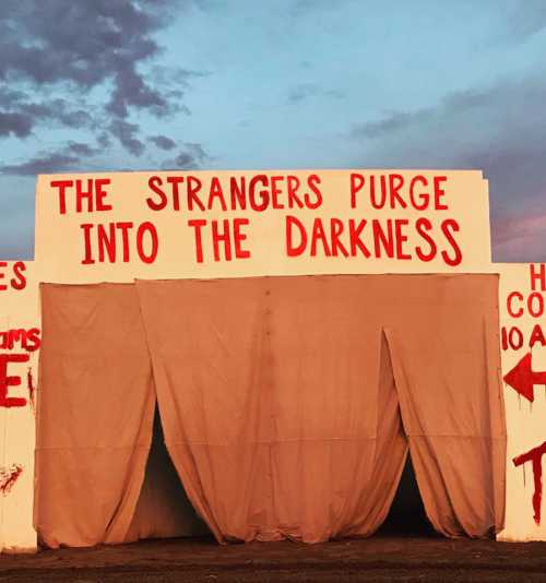 A sign reading "THE STRANGERS PURGE INTO THE DARKNESS" with a curtain entrance, set against a colorful sky.