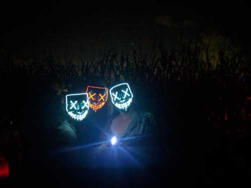 Three people wearing glowing skull masks stand in a dark field, holding a flashlight.