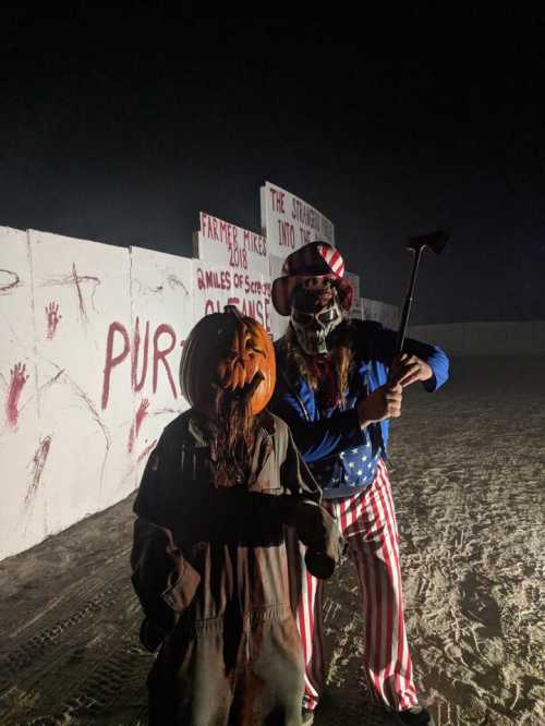 Two costumed figures stand in a foggy setting: one with a pumpkin head and the other in patriotic attire holding an axe.