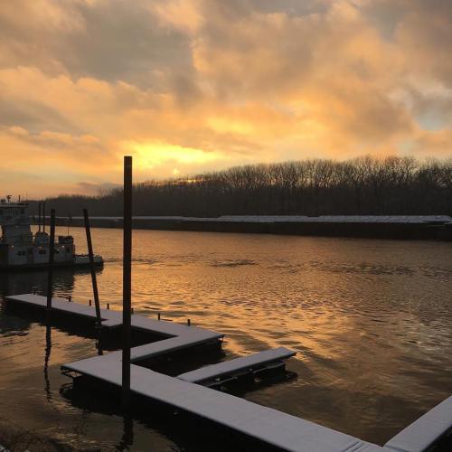 A serene river scene at sunset, with boats docked and trees silhouetted against a colorful sky.