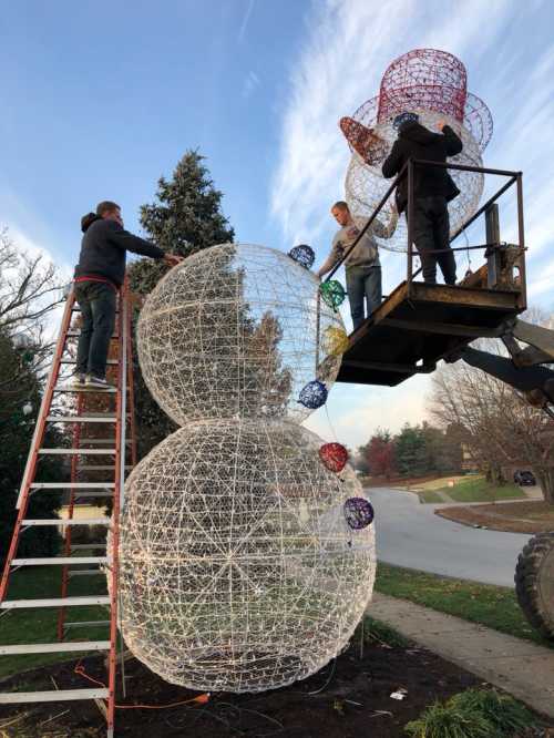 Workers assembling large, illuminated holiday decorations on a tree using a lift and ladder in a suburban area.