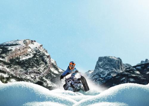 A snowmobiler jumps over a snowy landscape with mountains in the background under a clear blue sky.