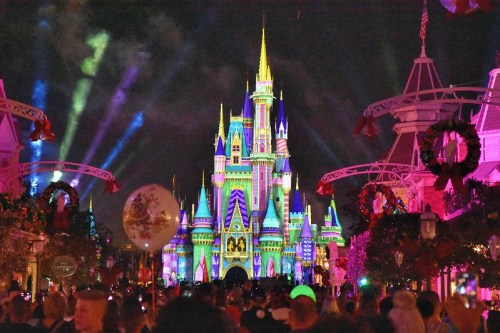 Colorful lights illuminate a castle at night, surrounded by festive decorations and a crowd of people.