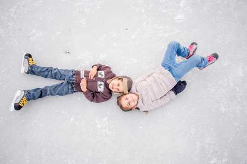 Two children lie on an icy surface, smiling and relaxed, wearing winter clothing and colorful shoes.