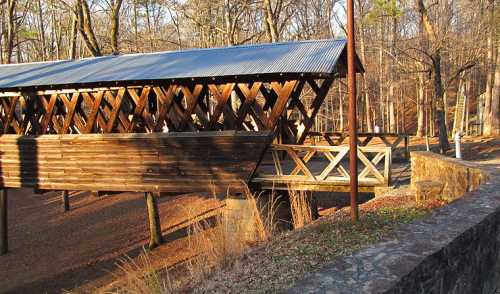 A wooden covered bridge with a metal roof, surrounded by trees and a gravel path in a wooded area.