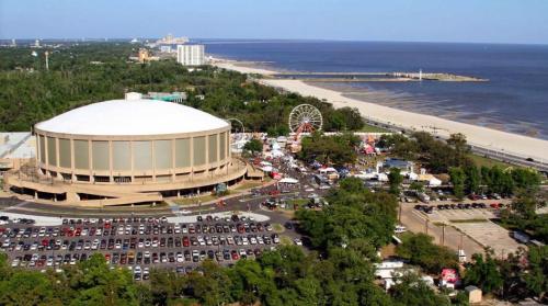 Aerial view of a large circular building near a beach, with a carnival and parking lot visible in the foreground.