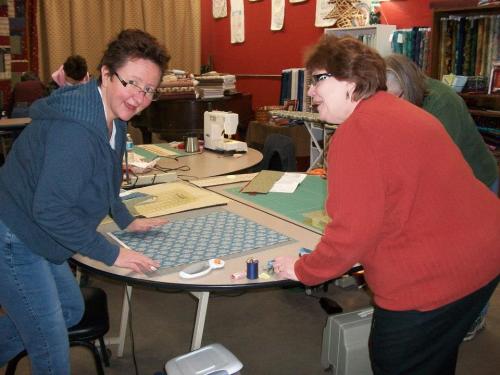 Two women work together at a table, cutting fabric and sewing supplies in a craft room filled with colorful materials.