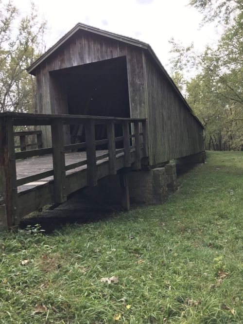 A rustic wooden covered bridge with a ramp, surrounded by greenery and trees.