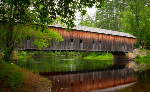 A rustic covered bridge spans a calm river, surrounded by lush greenery and reflected in the water below.