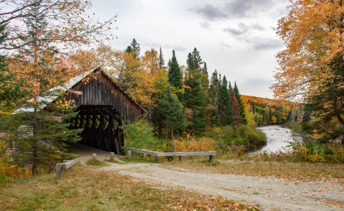 A rustic covered bridge surrounded by vibrant autumn foliage and a winding river under a cloudy sky.