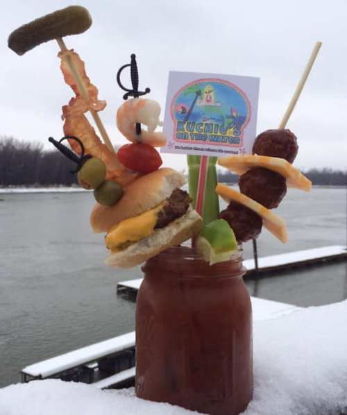 A colorful drink garnished with a mini burger, shrimp, pickles, and meatballs, set against a snowy riverside backdrop.