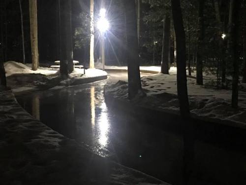 A serene nighttime scene of a snowy path beside a calm, reflective waterway illuminated by a bright light.