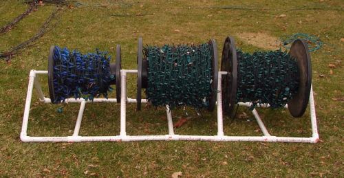 Three large spools holding colorful string lights, supported by a white PVC frame, on a grassy surface.