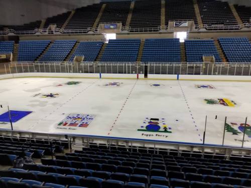 An empty ice rink with colorful painted designs on the ice, surrounded by blue seating in a large arena.