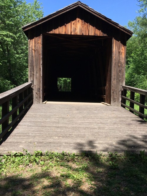 A wooden covered bridge surrounded by greenery, leading into a dark interior with sunlight filtering through.