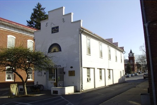 A white building with a sun emblem, labeled "Library," surrounded by trees and brick structures on a sunny day.