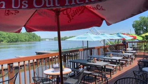Outdoor dining area with tables under colorful umbrellas, overlooking a calm river and green trees.