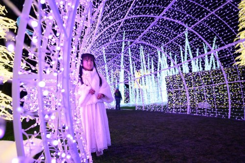 A child in a white dress stands inside a glowing archway of purple and white lights in a festive display.