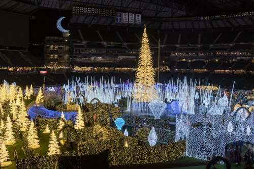 A festive display of colorful lights and decorations, featuring a tall illuminated tree in a stadium setting.