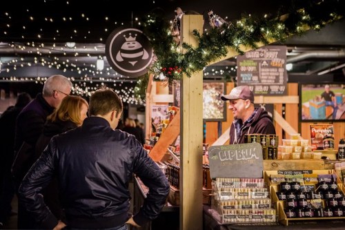 A festive market scene with people browsing a vendor's stall, decorated with lights and holiday decor.