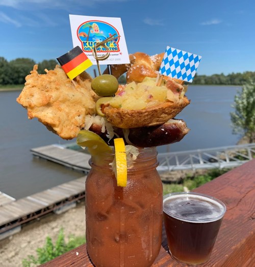 A large, colorful drink garnished with pretzels, olives, and flags, set against a river backdrop.