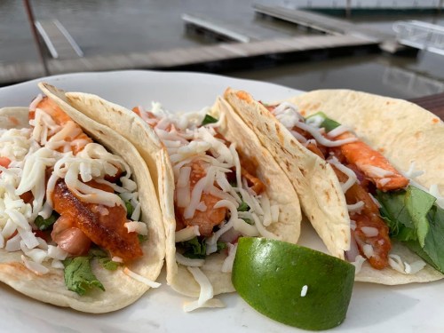 Three tacos filled with shredded cheese, lettuce, and fish, served with a lime wedge, by a waterfront.