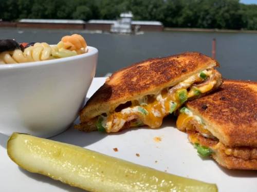 A grilled cheese sandwich with melted cheese and vegetables, served with a side of pasta salad and a pickle, by a river.