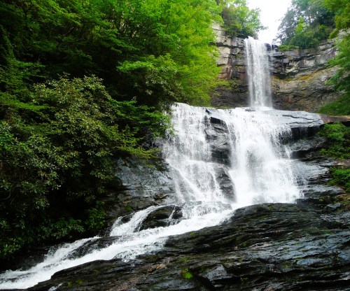 A cascading waterfall flows over rocky cliffs, surrounded by lush green trees and foliage.