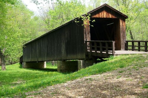 A wooden covered bridge stands on stone supports, surrounded by lush green trees and grass.
