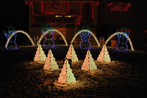 Colorful holiday lights display featuring snowmen and illuminated Christmas trees in a snowy yard.