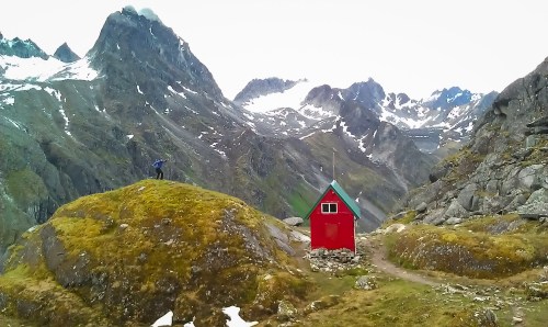 A small red cabin on a grassy hill surrounded by rugged mountains and snow-capped peaks under a cloudy sky.
