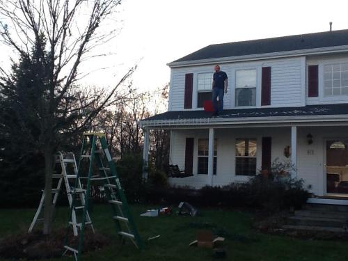 A person stands on a roof, holding a red bucket, with a ladder and tools nearby in a residential yard.