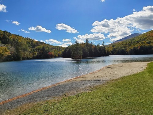 A serene lake surrounded by trees and mountains, with a clear blue sky and fluffy clouds reflecting on the water.