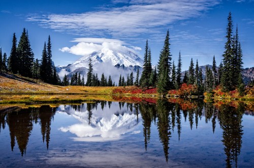 A serene landscape featuring a snow-capped mountain reflected in a calm lake, surrounded by evergreen trees and colorful foliage.