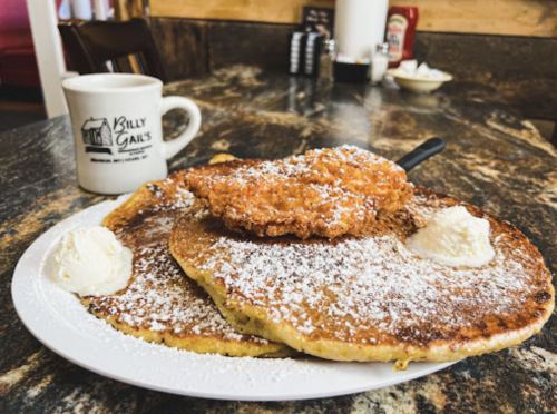 A plate of fluffy pancakes topped with fried chicken and dollops of butter, served with a coffee mug in the background.