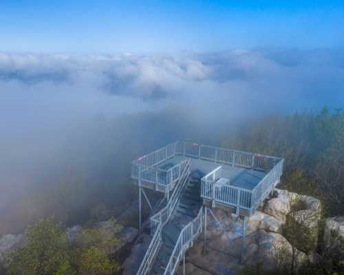 A viewing platform atop a rocky outcrop, surrounded by misty clouds and greenery under a clear blue sky.