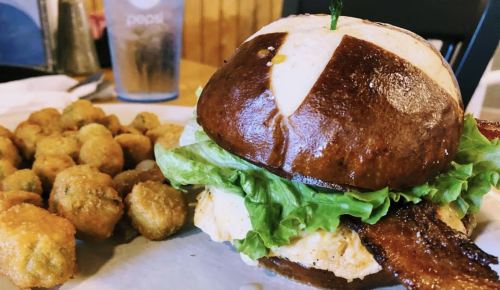 A close-up of a pretzel bun sandwich with lettuce and bacon, served with a side of fried okra.