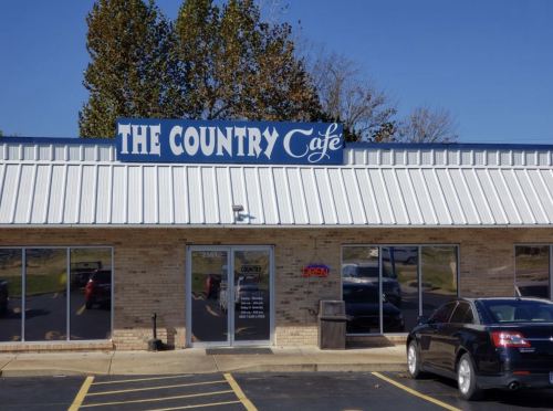 Exterior of The Country Café, featuring a blue sign, open door, and a parked car in front. Clear blue sky above.