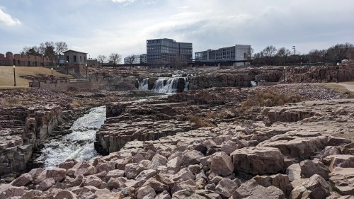 A scenic view of a waterfall cascading over rocky terrain, with modern buildings in the background under a cloudy sky.