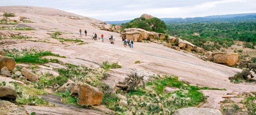 A group of hikers exploring a rocky landscape with sparse vegetation and distant hills under a cloudy sky.